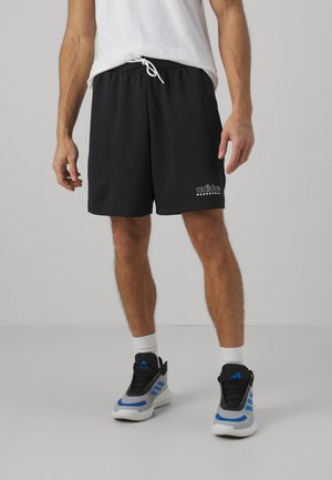 Homme portant un short de basket Adidas noir, des chaussettes blanches et des baskets Adidas gris-bleu, debout sur un sol gris clair uni.