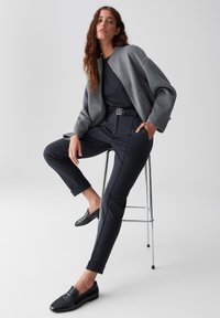 Woman with long wavy hair wearing a gray jacket, black top, dark cuffed pants, and black loafers sitting on a chrome stool against a white background.