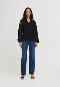Blusa nera con maniche in pizzo, design a V, abbinata a jeans larghi blu e sandali neri. Linee pulite e dettagli texture.