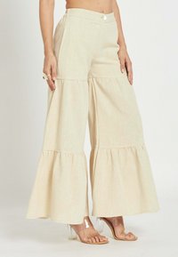 Pantalones beige de pierna ancha en capas con cintura de botones, combinados con sandalias transparentes de tacón alto, mostrando el abdomen y manos con anillos.