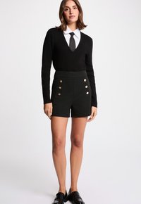 Pull noir à col en V avec manches longues associé à un short noir taille haute doté de boutons dorés. Le mannequin porte des mocassins noirs en cuir verni.