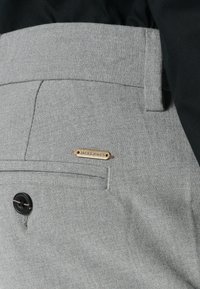Gros plan sur la poche arrière d'un pantalon gris avec un bouton noir et une petite étiquette en métal gravée "JACK & JONES" attachée au-dessus du bord de la poche.