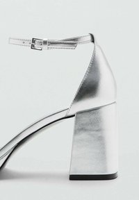 Strieborné sandále na vysokom podpätku s širokým blokovým podpätkom, s remienkom okolo členka a hladkým metalickým povrchom. Majú špicatú špičku a minimalistický dizajn.