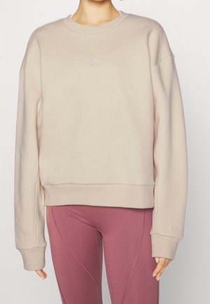 Frau trägt beige Adidas-Sweatshirt mit kleinem Logo und pinken eng anliegenden Leggings, steht vor einfachem hellem Hintergrund.