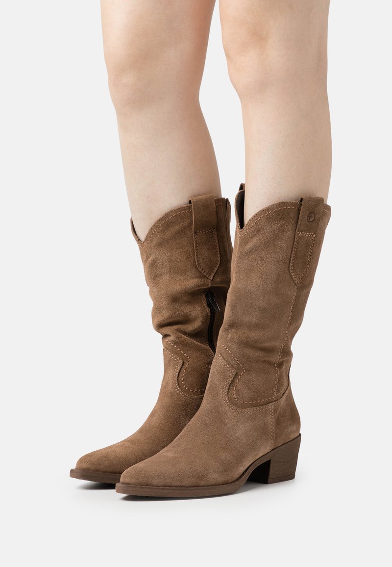 Cizme de cowboy din suede maro, cu vârf ascuțit, cusături decorative și toc stratificat. Au fermoar lateral și un design cu guler pliat.