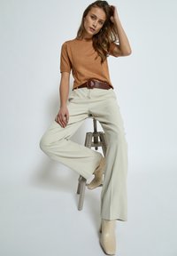 Hnědý pletený top s krátkým rukávem, krémové kalhoty s vysokým pasem, hnědý texturovaný opasek a béžové boty na podpatku, sedící na šedém stoličce.