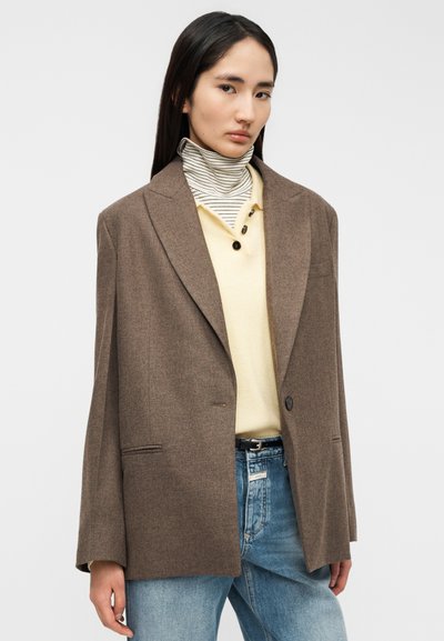 Blazer de lana marrón con cuello de muesca, cierre de un solo botón y dos bolsillos frontales, llevado sobre una camisa amarilla y un bufanda tubular a rayas.