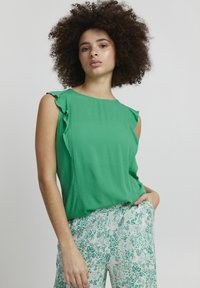 Top verde senza maniche con dettagli arricciati sulle spalle, realizzato in tessuto testurizzato. Abbinato a pantaloni bianchi con un motivo floreale verde.