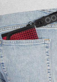 Red and black patterned pouch with a zip closure partially tucked into a light blue denim pocket. Black strap attached to the pouch.
