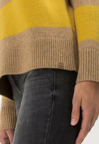 Striktrøje med skiftende gule og tan-striber, med ribbet kant og manchetter, parret med mørkegrå jeans. Diskret logo mærke synligt.