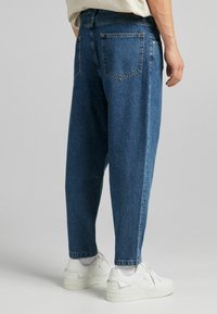 Blue denim jeans with a relaxed fit, visible back pockets, and a tapered ankle. Worn with white sneakers and ribbed cuffs.
