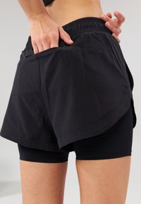 Černé sportovní šortky s elastickým pasem, které mají volnou vnější vrstvu přes upnuté vnitřní šortky. Hladká textura a minimalistický design.