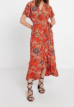 Femme portant une robe portefeuille à fleurs orange vif avec des manches volantées et des sandales à talons noires à fines brides, debout devant un fond uni.