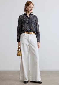 Camisa azul-marinho com padrão floral e botões, calças brancas de perna larga, cinto dourado, pequena mala amarela e sapatos pretos; tecido suave e cores contrastantes.
