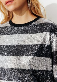 Pull à paillettes rayé avec des rayures horizontales noires et argentées, un col côtelé noir et une texture lisse et scintillante.