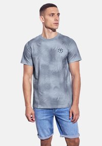 Šedé tričko s krátkým rukávem s texturovaným vzorem, s malým černým grafickým designem na levé straně hrudi. Nosené s lehkými denimovými šortkami.