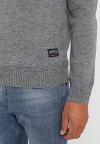 Närbild av en person som bär en grå stickad tröja med en liten svart etikett och blå denimjeans, med handen avslappnad vid sin sida.