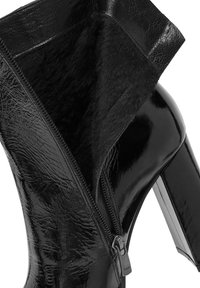 Botín de tobillo de charol negro con un acabado brillante, cierre de cremallera y un tacón alto de bloque. Interior forrado con material suave en negro.