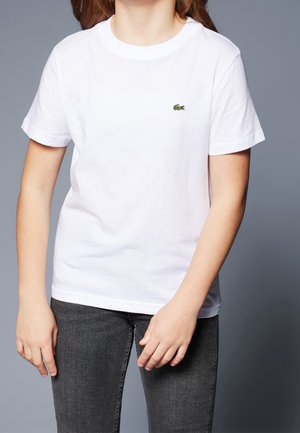 Person med en ensfarvet hvid kortærmet t-shirt med et lille grønt krokodillelogo på brystet og mørkegrå jeans mod en grå baggrund.