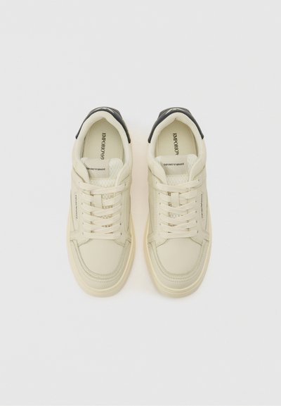 Beige leather sneakers with a textured white mesh tongue and a black heel accent. Features round toe, flat laces, and minimalistic design.