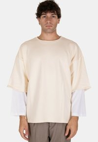 T-shirt oversize a maniche corte di colore crema, realizzato in tessuto morbido, con colletto rotondo e maniche lunghe bianche sottostrato.