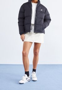 Osoba nosząca czarną kurtkę puchową, białą bluzkę, białą mini spódnicę, czarne skarpetki i białe sneakersy stoi na niebieskiej podłodze przy białej ścianie.