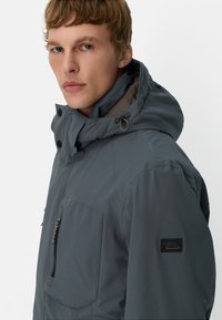 Grijze jas met capuchon, voorzien van een verticale ritszak aan de voorkant en zijzakken. Gladde textuur, minimale stiksels en een logo-opdruk op de mouw.