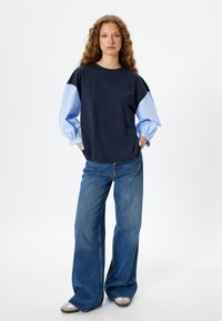 Marineblaue Baumwoll-T-Shirt mit hellblauen gestreiften Puffärmeln, kombiniert mit hellblauen weiten Jeans und grauen Sneakers, vor einem schlichten Hintergrund stehend.