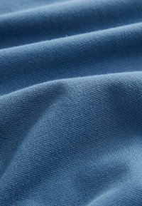 Sininen kangas, jossa on teksturoitu pinta, hienovarainen neulekuvio ja lempeät taitteet, jotka antavat sille sileän ja pehmeän ilmeen.