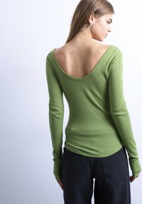 Νεαρή γυναίκα που φοράει πράσινο μακρυμάνικο τοπ με βαθύ κόψιμο στην πλάτη και μαύρο παντελόνι, γυρισμένη προς τα πίσω μπροστά σε απλό λευκό φόντο.