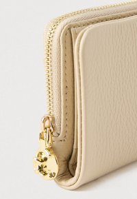 Beige leren portemonnee met textuur, gouden rits en ritsring in klaverbladvorm, weergegeven in close-up op een lichte, effen achtergrond.