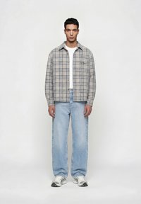 Νέος άνδρας στέκεται όρθιος, φοράει γκρι καρό πουκάμισο με κουμπιά πάνω από λευκή μπλούζα, ανοιχτό μπλε τζιν με φαρδιά παντελόνια και λευκά αθλητικά παπούτσια, σε απλό λευκό φόντο.