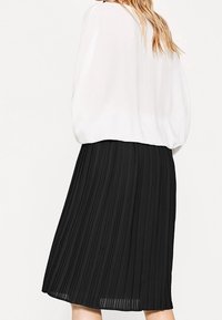 Gonna nera plissettata con un design fluido e testurizzato, abbinata a una blusa bianca larga. La gonna presenta pieghe verticali per una maggiore dimensione.