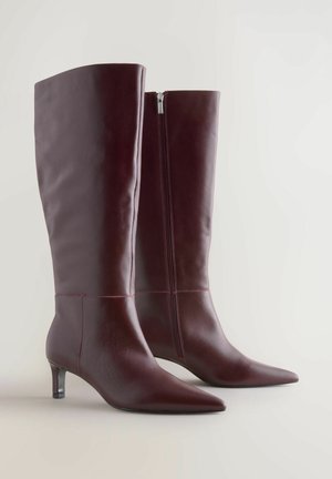 Burgundy nahka polvipituiset saappaat, joissa on kapea kärki, koristeellinen ompelu ja sivuvetoketju. Varustettu matalalla, hoikalla korolla.