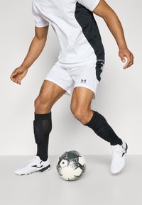 Weißes Sportshirt mit schwarzem Seitenpanel, weiße Shorts, schwarze Kompressionssocken und weiße Fußballschuhe neben einem schwarz-weißen Fußball.