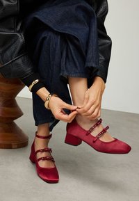 Chaussures à talons hauts en satin rouge avec un bout carré, arborant trois brides à boucle et un talon épais, portées par une personne ajustant la bride.