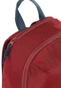 Esquina de una mochila roja de nailon, con un asa de tela azul oscura y un tirador de cremallera. Superficie texturizada con detalle de costuras visibles.