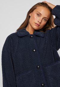 Jauna moteris su šviesiai rudais plaukais, vilkinti tamsiai mėlyną tekstūruotą mygtukų paltą, pakėlusi vieną ranką, kad paliestų savo plaukus.