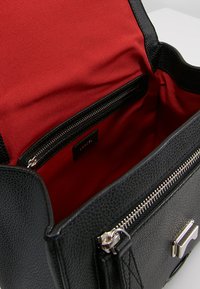 Sac à main en cuir noir avec une finition texturée. Doublure en tissu rouge à l'intérieur, comportant une poche zippée et une étiquette de marque. Accents en métal argenté.