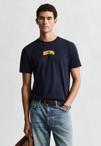 Camiseta de algodón azul marino con cuello redondo, con el logo amarillo bordado "GANT 1949" en el pecho. Se lleva con jeans azul claro.