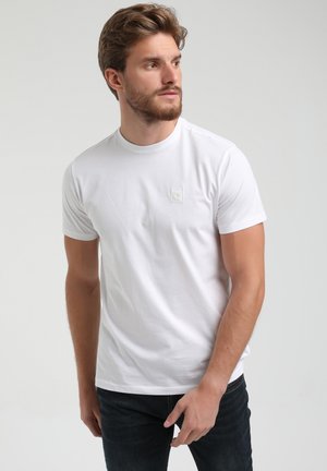 T-shirt en coton blanc avec un col rond et des manches courtes, arborant un petit patch logo sur la poitrine gauche. Texture lisse.