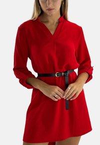 Robe rouge à col, manches longues, poignets boutonnés et tissu texturé. Une ceinture noire avec une boucle dorée serre la taille.