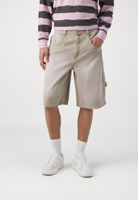 Beige denimshorts med avslappnad passform, framfickor och fransad fåll. I kombination med en långärmad skjorta i rosa och svart randigt mönster samt vita sneakers.