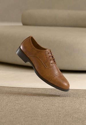 Zapato brogue de cuero marrón con perforaciones decorativas, suela negra y cordones, colocado sobre una alfombra beige y cerca de un sofá beige.