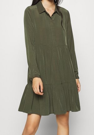 Nainen pukeutuneena oliivinvihreään pitkähihaiseen paitamekkoon, jossa on kaulus ja kerroksittain laskostettu hame, seisoo yksinkertaisen taustan edessä.