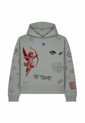 Blondine vrouw met een grijze hoodie met rode cupido, ogen en de tekst "Je T'aime", gecombineerd met zwarte cargobroeken en witte sneakers, staand.