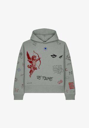 Femme blonde portant un sweat à capuche gris avec un cupidon rouge, des yeux et le texte « Je T'aime », associée à un pantalon cargo noir et des baskets blanches, debout.