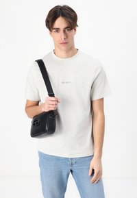 Black crossbody bag made of smooth synthetic material, featuring a raised logo on the front, carried on a wide adjustable strap.