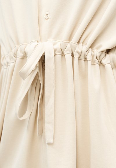 Μπεζ βαμβακερό ύφασμα με ελαστική ζώνη στη μέση, μαζεμένη και δεμένη με φιόγκο, παρουσιάζοντας λεπτομέρειες με κουμπιά πάνω από τη μέση.