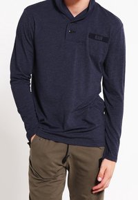 Marinblus med lång ärm och sjalkrage, försedda med en knapp och en bröstficka med texten "RAW" i mörkt tyg.